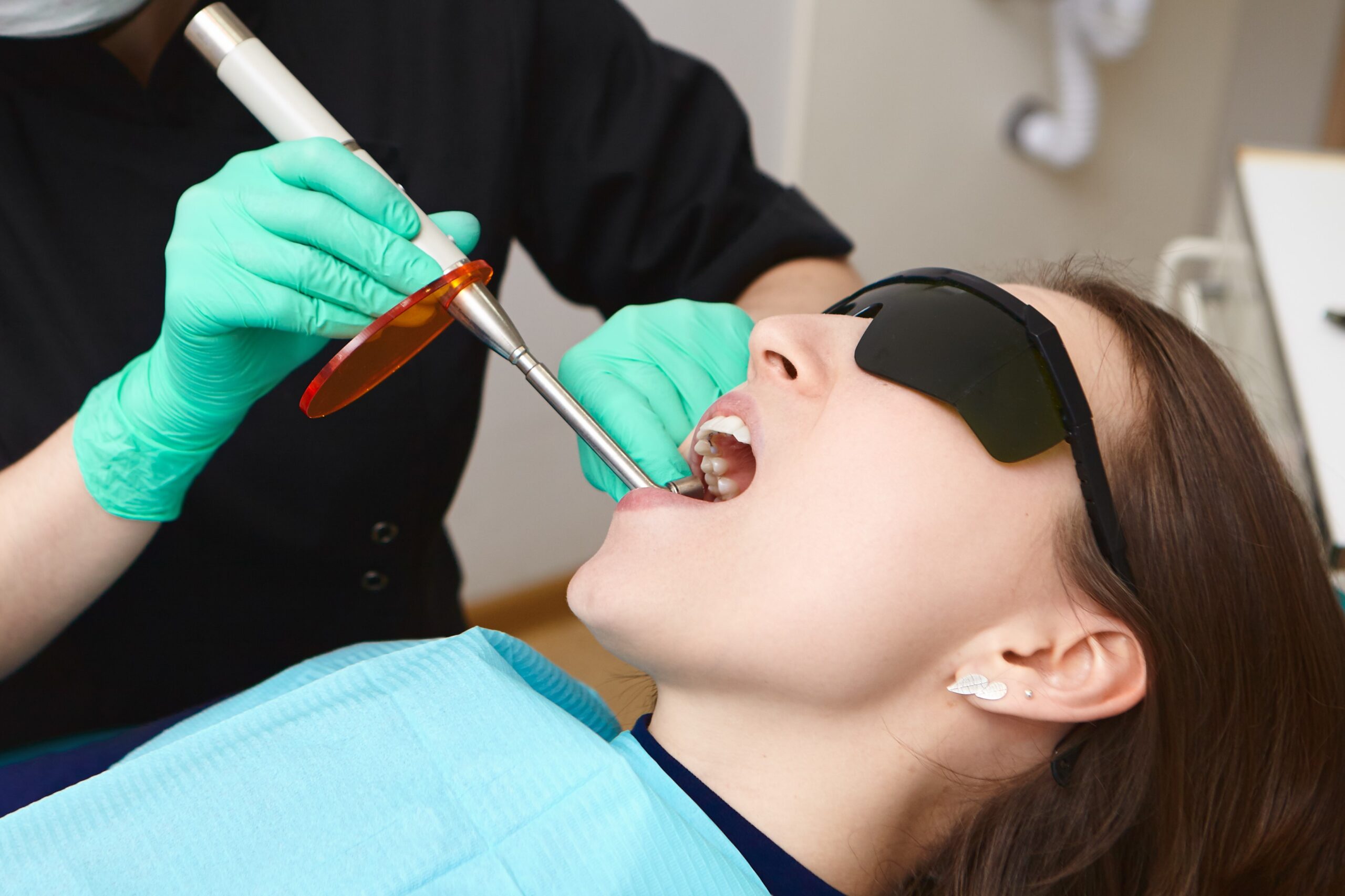 People, healthcare, modern technology, innovations and stomatology concept. Picture of woman patient in black goggles getting her teeth treated by female hygienist using dental curing light